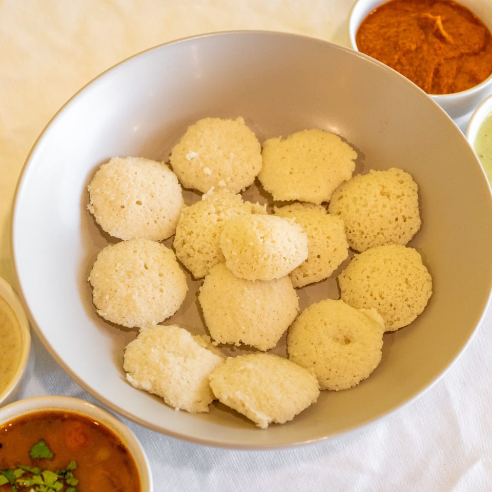 A bowl of fluffy idli, with small bowls of red and green chutneys and sambar.