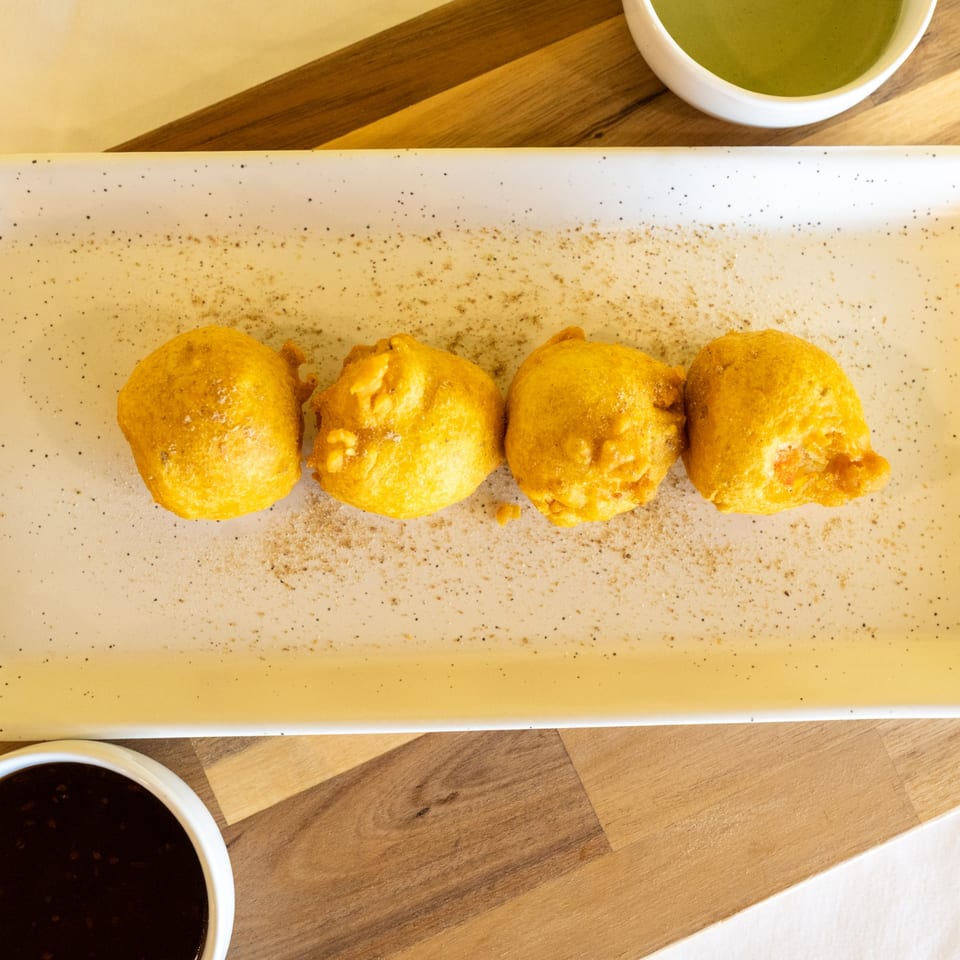 Four golden-brown fried fritters dusted with spices on a white rectangular plate, with two dipping sauces.
