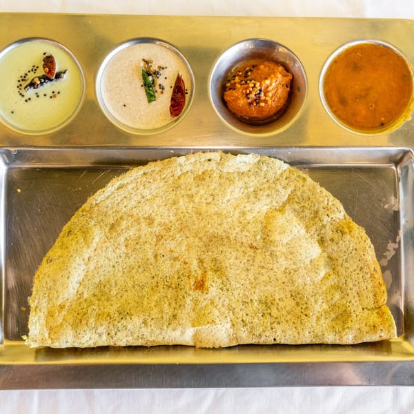 A large dosa with four small bowls of chutneys and sambar on a metal tray.