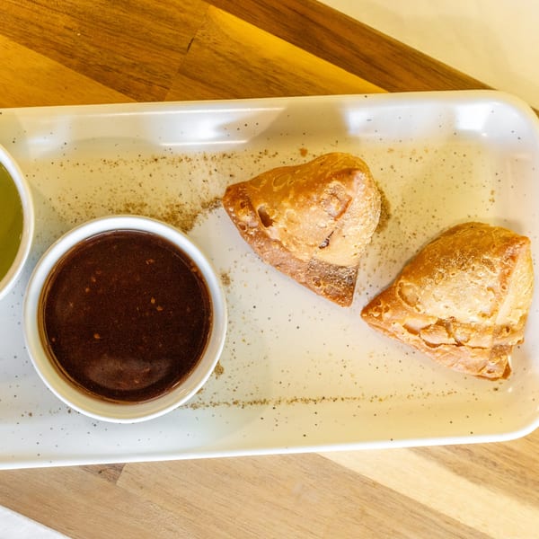 Two golden-brown samosas and two dipping sauces on a white rectangular plate.