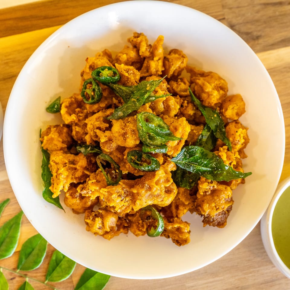 Crispy fried chicken pieces, garnished with green chili slices and curry leaves, in a white bowl.