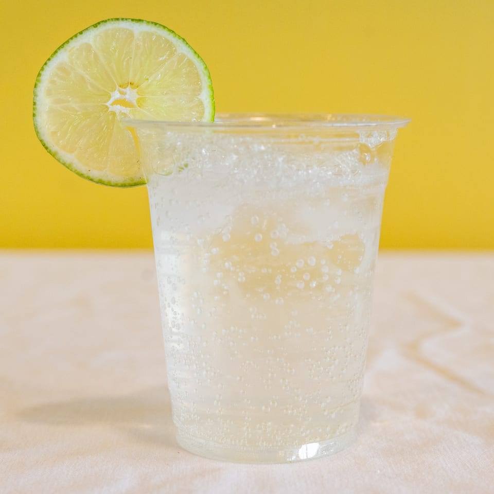 Sparkling clear beverage in a plastic cup, garnished with a fresh lime slice, against a yellow background.