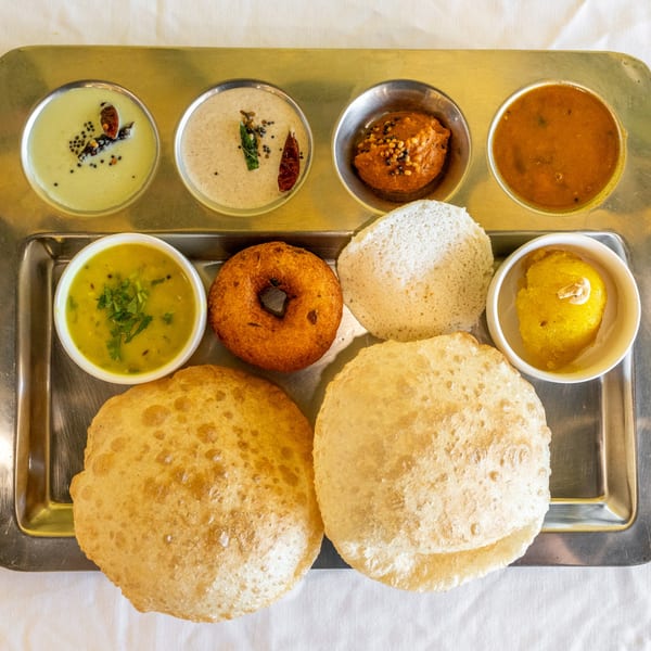 South Indian thali with puri, vada, idli, sambar, dal, and chutneys on a metal tray.