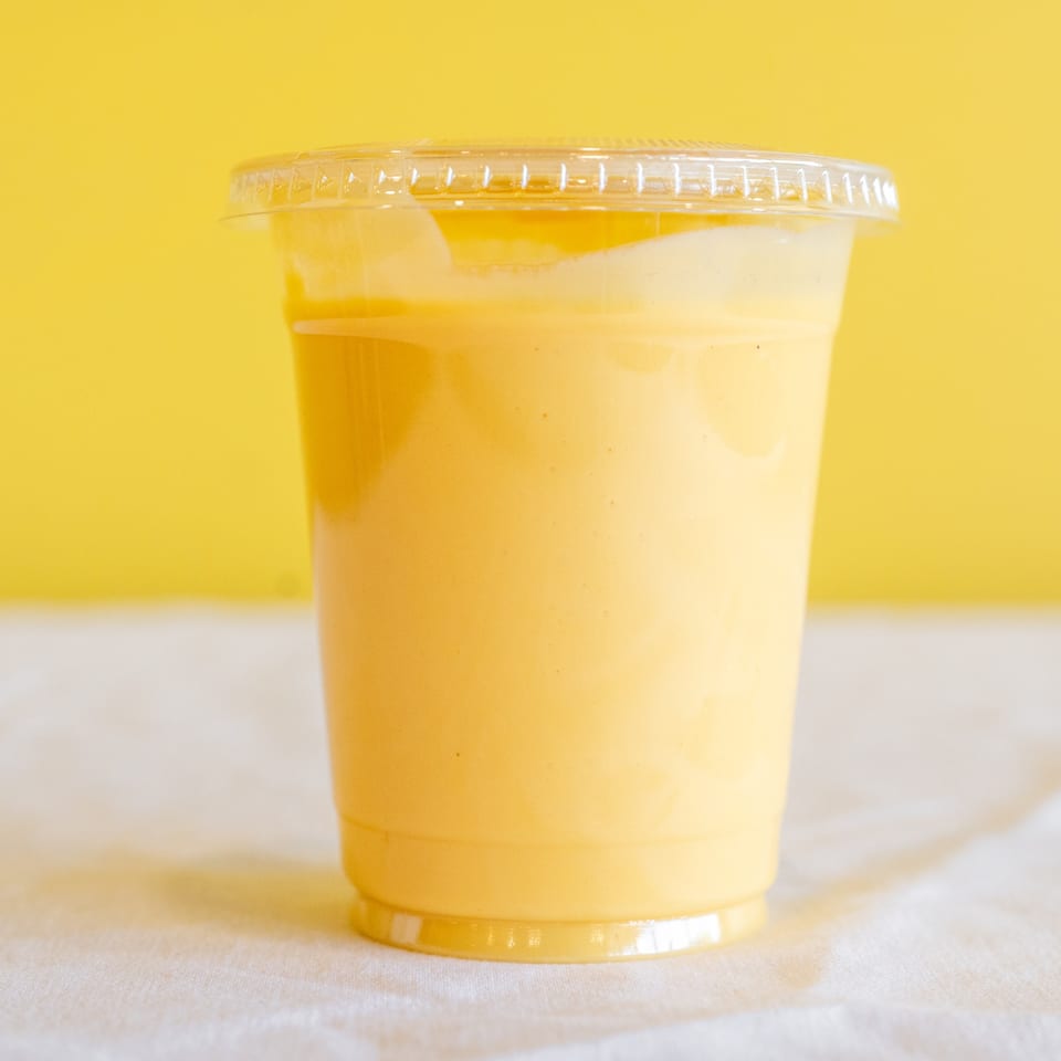 A vibrant yellow mango lassi in a clear plastic cup with a lid, against a yellow background.