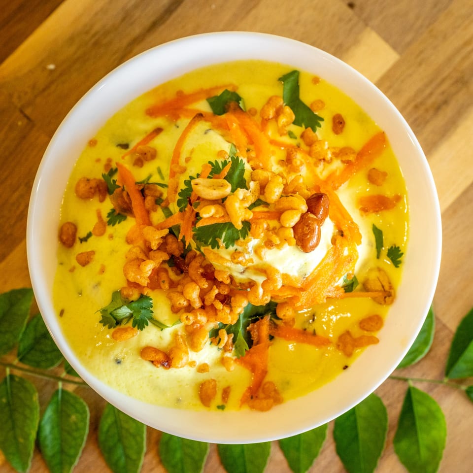 Overhead view of a yellow soup topped with shredded carrots, peanuts, and cilantro in a white bowl.