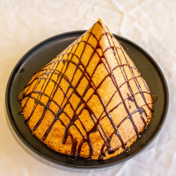 A golden-brown cone dosa, drizzled with a crisscross pattern of chocolate syrup, on a black plate.